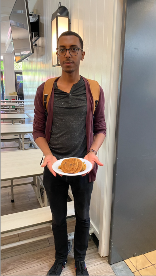 Iskindir is holding a plate of cookies in the San Domenico cafeteria and dressing on trend with a maroon cardigan, black jeans and casual sneakers.